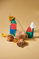 A contemporary still life with a cup of cocoa, wooden cubes, and dried maple leaves on a plain beige background.