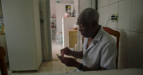 One Elderly Black man sitting at dining table in a cozy kitchen, concentrating on a smartphone, capturing the essence of daily routine and technology in a relaxed home environment