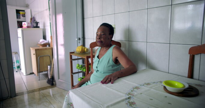 Elderly African American woman sitting quietly at the dining table in a modest home kitchen, hand resting on the table, reflecting solitude and quiet moments of introspection