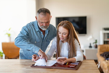 Fototapeta premium Elderly man reviews documents with younger woman in bright living room while she focuses on her phone
