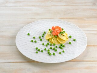 A serving of pasta is garnished with cooked shrimp and a scattering of bright green peas. The presentation is elegant, set against a pale, neutral backdrop