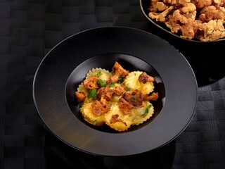 A close-up shows ravioli topped with sauteed chanterelle mushrooms. The food is served in a black bowl against a dark backdrop. Fresh parsley adds a touch of green