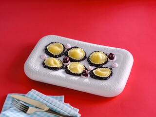 Six ravioli with golden pasta and a dark fruit reduction are presented on a white plate with dollops of pink cream, a knife, a fork, and a blue-checkered napkin complete the setting