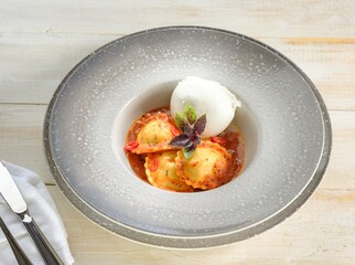Three ravioli are served in a vibrant red sauce, adorned with fresh basil and a ball of mozzarella. This culinary creation rests on a stylish gray plate, atop a wooden surface