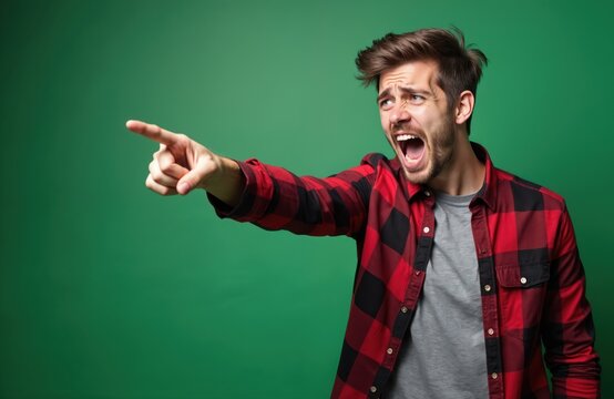 Young man in red plaid shirt yells and points forward. Angry male shouts, expresses frustration. Expressing outrage, anger, or giving strong direction. Studio portrait. Copy space.