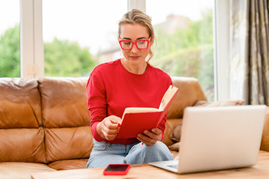 Woman reading a book while sitting on a sofa next to a laptop in a cozy living room during the afternoon