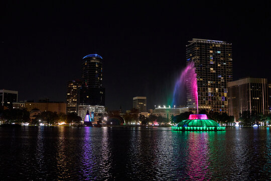 A wide city panorama shows a glowing fountain and colorful lights across the water. Towers and high-rises shine in a calm night scene.