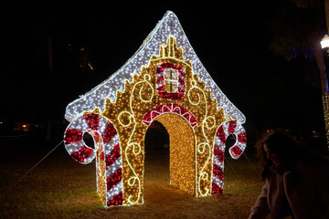 Naklejka premium A large gingerbread house arch made of LED lights glows at night. Festive candy cane columns and bright decoration welcome families in a winter park.
