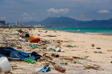 A beach is strewn with various types of waste, including plastic bottles and containers. The clear blue water contrasts sharply with the litter. Mountains loom in the background