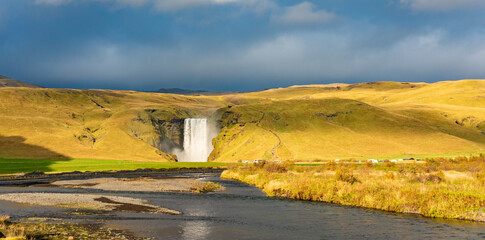 Skogafoss waterfall in Iceland cascades down a steep cliff, creating a powerful mist that forms a vibrant rainbow. The surrounding lush green landscape enhances its majestic beauty © Kotangens