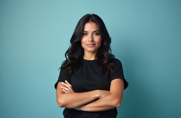 Obraz premium Young hispanic woman with dark wavy hair wears black t shirt, arms crossed. Confident pose against blue background. Direct look, neutral expression.