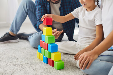 Family playing at home, child stacking colorful blocks with help of parents sitting on carpet of...