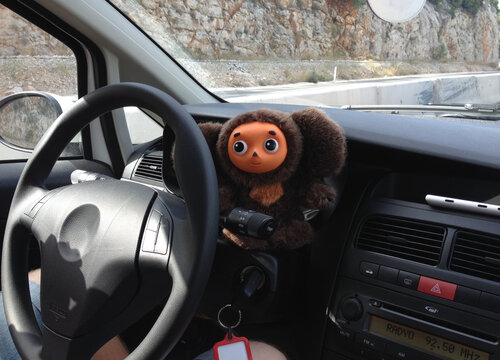 Plush toy Cheburashka sitting by the steering wheel inside a car. Travel, personality, and lighthearted company on a scenic journey.