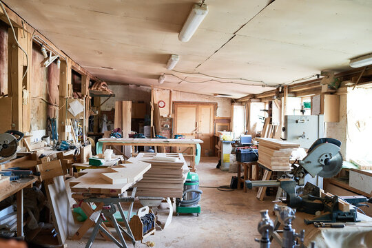 Empty woodworking workshop showing stacks of cut lumber, power tools, workbenches, and sawdust covering floor, natural light entering through windows, no people visible