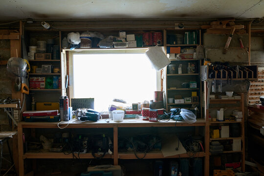 Wooden workbench covered with assorted tools, power drills, extension cords and containers, shelves filled with hardware supplies and equipment surrounding window in cluttered workshop