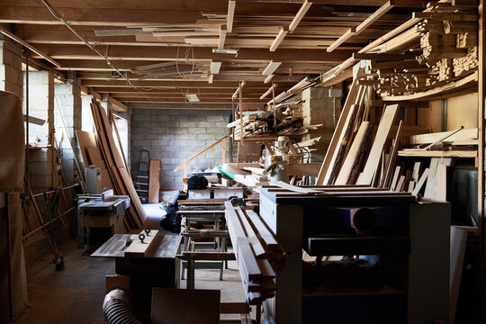Empty woodworking workshop showing stacks of lumber, woodworking machines, and scattered tools in spacious industrial setting, sunlight streaming through windows illuminating workspace