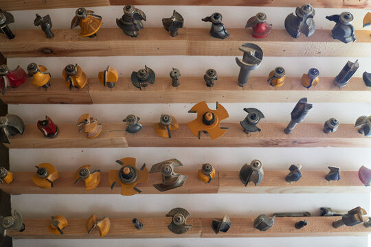 Assorted woodworking router bits arranged on wooden rack, various shapes and sizes organized in rows, metal tools used for shaping, cutting, and carving wood surfaces