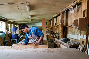Young adult Caucasian man working in carpentry workshop using hand tool on wooden plank, focusing on craftsmanship with wood shavings scattered on large workbench surface