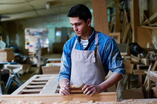 Young adult Caucasian man working in carpentry workshop using hand planer on wooden plank, focused expression, surrounded by woodworking tools and unfinished furniture