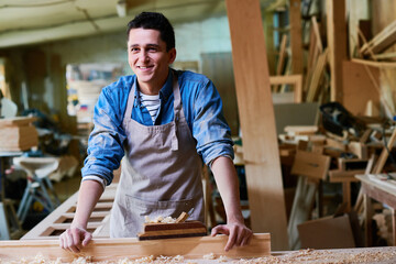 Caucasian young adult man standing at woodworking bench smiling while working in carpentry workshop, hands resting on table surrounded by wood shavings and tools