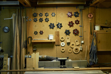 Woodworking workshop showing various circular saw blades and wooden discs mounted on wall, wooden planks and boards stacked around workspace, industrial equipment visible