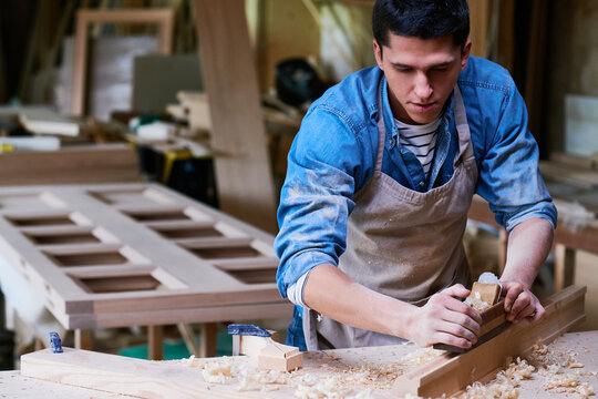 Young adult Caucasian man working with wood planer, shaping wooden plank in carpentry workshop, focused on craftsmanship, wood shavings scattered on workbench, wearing apron