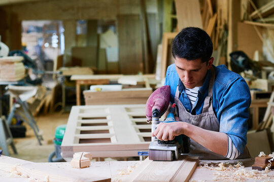 Young adult Caucasian man using electric sander while working on wooden board in carpentry workshop, focusing on precise craftsmanship with wood shavings scattered on workbench
