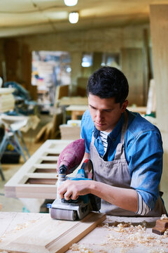 Young adult Caucasian man using electric sander on wooden board in carpentry workshop, focusing on smoothing surface while wearing apron, wood shavings scattered on workbench
