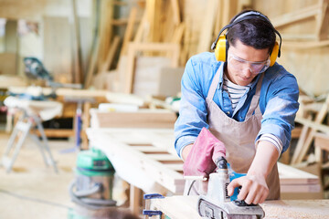Young adult Caucasian man wearing protective goggles and earmuffs using electric planer on wooden board in carpentry workshop, focusing on precise woodworking task