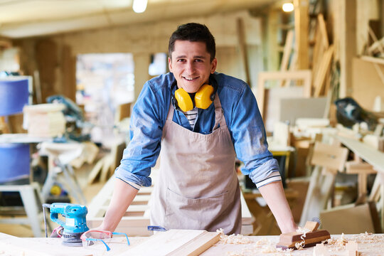 Portrait of young adult Caucasian man smiling and leaning on workbench in carpentry workshop, wearing apron and protective headphones, surrounded by woodworking tools and materials - Powered by Adobe