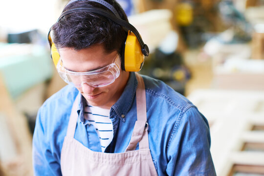 Young adult Caucasian man wearing protective goggles and earmuffs working in carpentry workshop, focusing on woodworking project with safety equipment, background showing blurred tools and materials