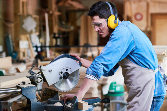 Young adult Caucasian man operating circular saw in woodworking workshop, wearing safety goggles and ear protection, focusing on precise cutting of wooden board, surrounded by carpentry tools