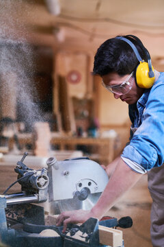 Young adult Caucasian man operating circular saw in woodworking workshop, wearing protective eyewear and ear protection, focusing on cutting wood with sawdust flying in air