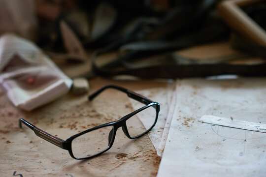 Eyeglasses resting on dusty workbench surrounded by scattered papers and tools, suggesting workspace environment with focus on vision correction accessory and creative process