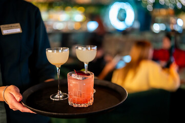Waiter's hands holding a black tray with three garnished cocktails, one pink and two pale yellow, in a dimly lit bar setting.