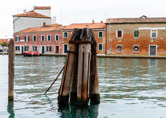 Murano, Italy - November 21, 2024: Scenic view of Murano island waterfront with wooden posts and colorful buildings reflecting in calm water, showcasing the charm of Venetian architecture
