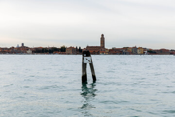 Murano, Italy - November 21, 2024: Scenic view of Murano Island with wooden post in foreground, showcasing tranquil waters and historic architecture in the background, ideal for travel enthusiasts