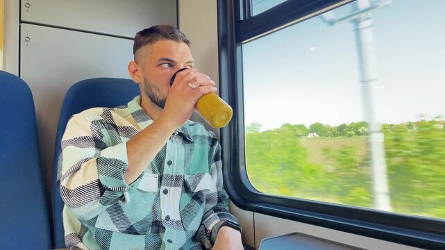Young man in checkered shirt holding yellow thermocup, drinking coffee and looking through train window at countryside view. 4k horizontal footage