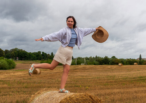 Cheerful woman in light clothes stands on a hay bale holding her hat in the wind. The playful pose and cloudy rural background express energy, joy, and carefree summer spirit.
