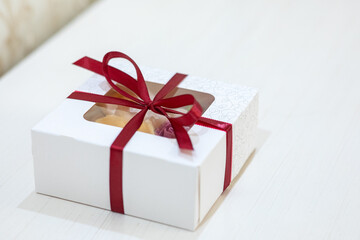 Small white cardboard gift box tied with a red satin ribbon, featuring a clear window and decorative pattern on a light surface
