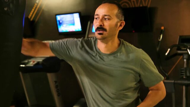 Slow motion video of an amateur man with mustache awkwardly hitting a punching bag. Man in his 30s with no experience trying to box and kick, shown in 4K slow motion.