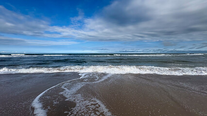 Image of a beach with a horizon separating the sea from the sky. Waves break on the shore, creating white foam that spreads across the wet sand, and the sky is partially light blue with clouds.