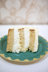 Single slice of layered vanilla sponge cake with rich white cream frosting on a teal and gold decorative paper plate