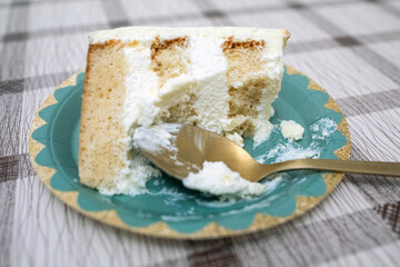 Slice of vanilla sponge cake with white cream frosting and a golden spoon on a decorative teal paper plate
