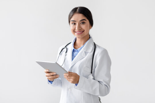 A cheerful millennial Indian lady doctor in a white coat stands against a white background. She holds a tablet and looks at the camera, promoting health care and remote consultations.