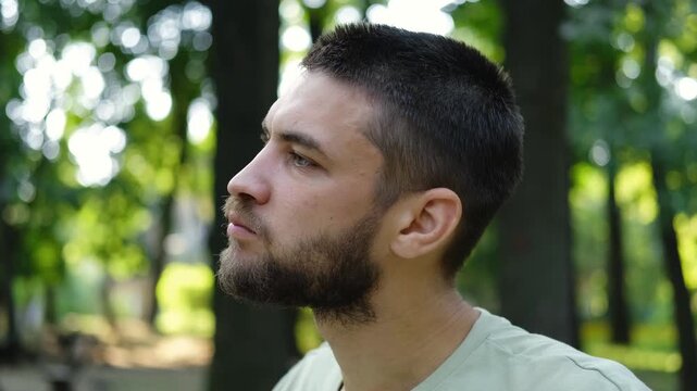 Young man in light green t shirt sitting on bench in park, eating red apple surrounded by green trees and sunlight. 4k horizontal footage