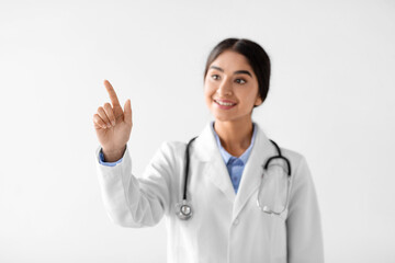 A smiling young Hindu woman doctor wearing a white coat and stethoscope points her finger to an...