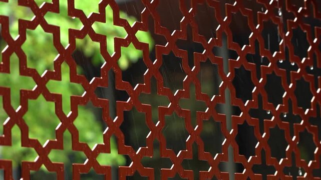 Shadowed rust lattice with green blur; textured metal grille casting patterned shadows, muted daylight and leafy bokeh behind, atmospheric garden barrier conveying privacy and aged