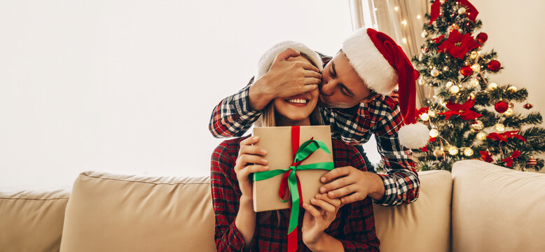 Christmas. Love. Presents. Man is covering his woman's eyes, kissing her in cheek and giving a gift box. Both are in Santa hats sitting at home near the Christmas tree