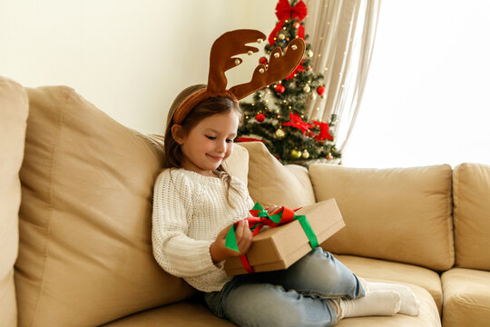 Happy little girl with reindeer antlers headband sits on a cozy beige sofa holding a Christmas gift box with red and green ribbon, smiling near a decorated Christmas tree.